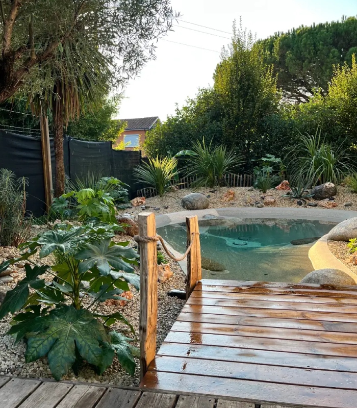 Aménagement extérieur autour d’une piscine lagon par CAP PAYSAGES Piscine lagon avec jardin paysager, végétation méditerranéenne et passerelle en bois